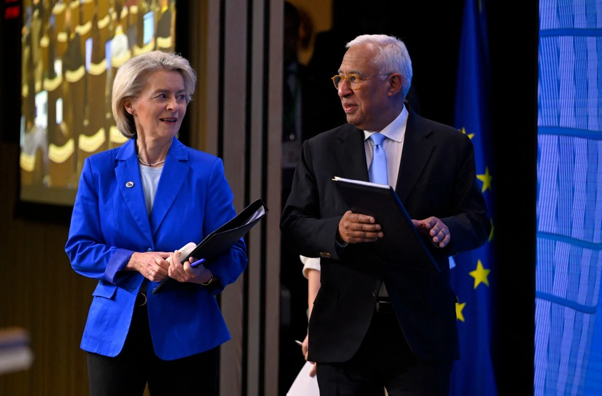 Przewodnicząca Komisji Europejskiej Ursula von der Leyen i przewodniczący Rady Europejskiej Antonio Costa Kobieta w niebieskiej marynarce oraz mężczyzna w garniturze rozmawiają pośród oficjalnej scenerii, oboje trzymają dokumenty, w tle widoczna flaga Unii Europejskiej.