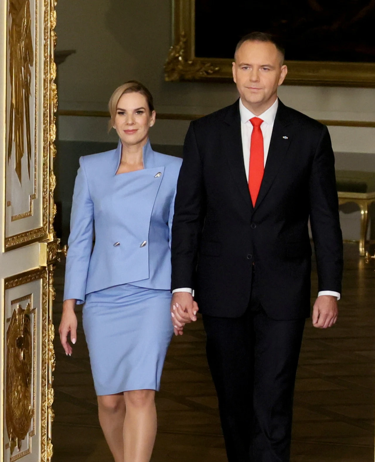Elegancka para trzymająca się za ręce, kobieta w niebieskim kostiumie i mężczyzna w garniturze z czerwonym krawatem, idą razem przez ozdobne, złocone wnętrze.