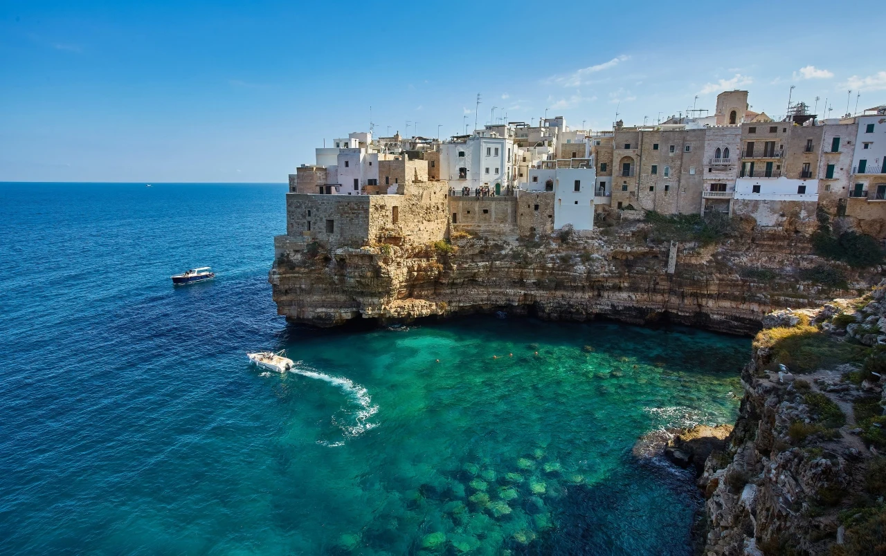 Polignano a Mare Białe budynki nad skalistym urwiskiem nad lazurowym morzem, z dwiema łodziami pływającymi w krystalicznie czystej wodzie.