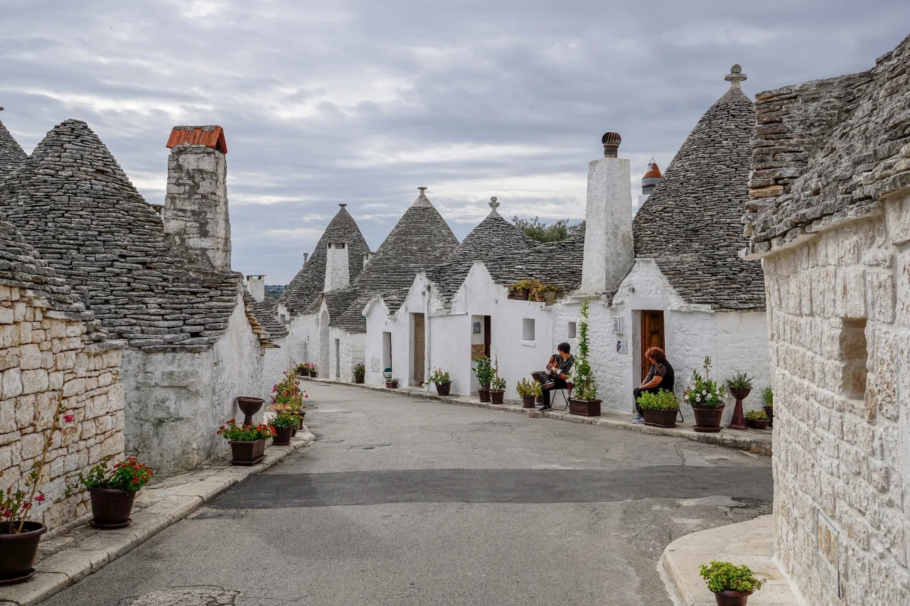 Alberobello kamienne domy o stożkowych dachach ułożonych z szarego łupka, ulica wyłożona asfaltem, liczne donice z zielonymi roślinami ustawione przy ścianach budynków, dwie osoby siedzące przy wejściu do jednego z domów