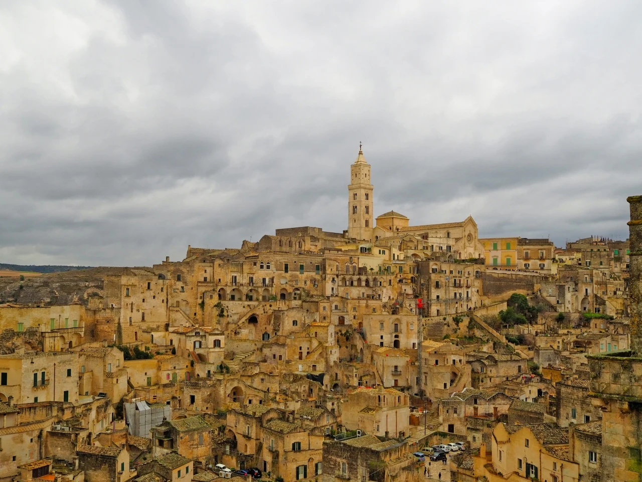 Matera gęsto zabudowane kamienne domy tworzące historyczne miasto na wzgórzu, zdominowane przez dużą kościołową wieżę, wszystko w ciepłych, piaskowych odcieniach pod pochmurnym niebem