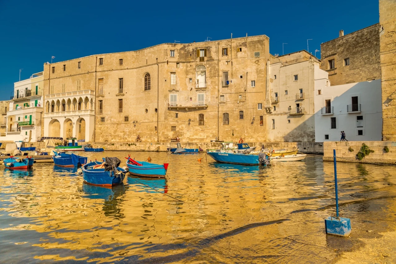 Monopoli Stare kamienne budynki nad wodą, kolorowe łódki zacumowane przy brzegu i odbicia architektury na spokojnej tafli portu w słonecznym świetle.