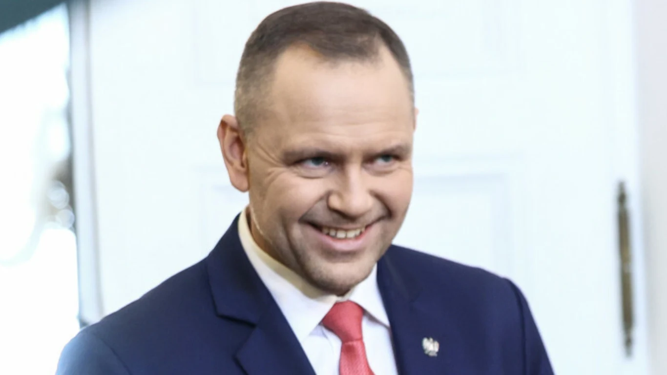 mężczyzna w garniturze z białą koszulą i czerwonym krawatem, uśmiechnięty, w tle jasne drzwi