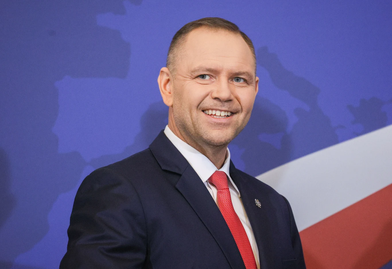 Karol Nawrocki Uśmiechnięty mężczyzna w garniturze i czerwonym krawacie na tle niebieskiej mapy z fragmentem biało-czerwonej flagi.