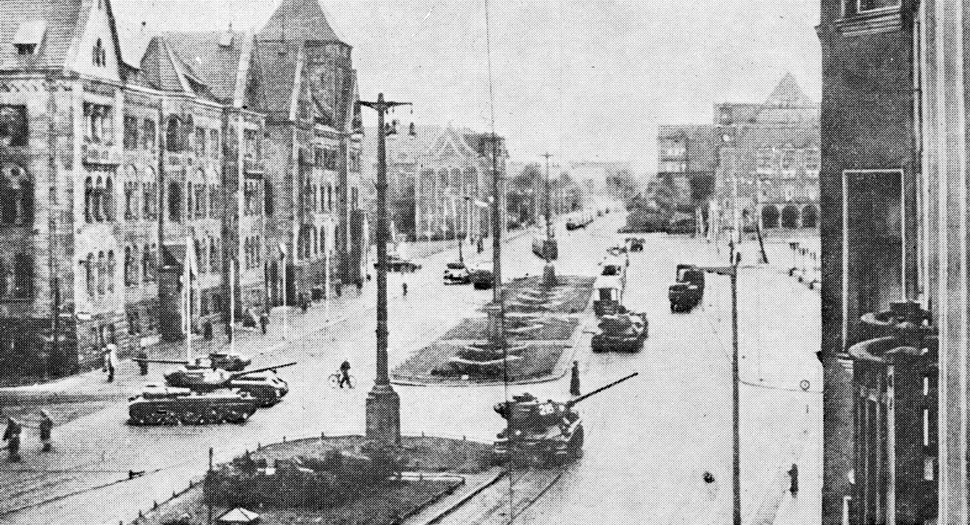kolumna czołgów poruszająca się szeroką ulicą wśród historycznych, wielopiętrowych budynków miejskich, pojedynczy piesi na chodnikach, pusta przestrzeń na środku jezdni