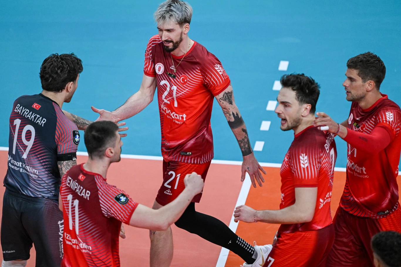 Grupa siatkarzy w czerwonych strojach świętuje punkt na boisku, jeden z zawodników wyciąga rękę w geście gratulacji, a pozostali są w ruchu i wykazują dużą emocjonalność.