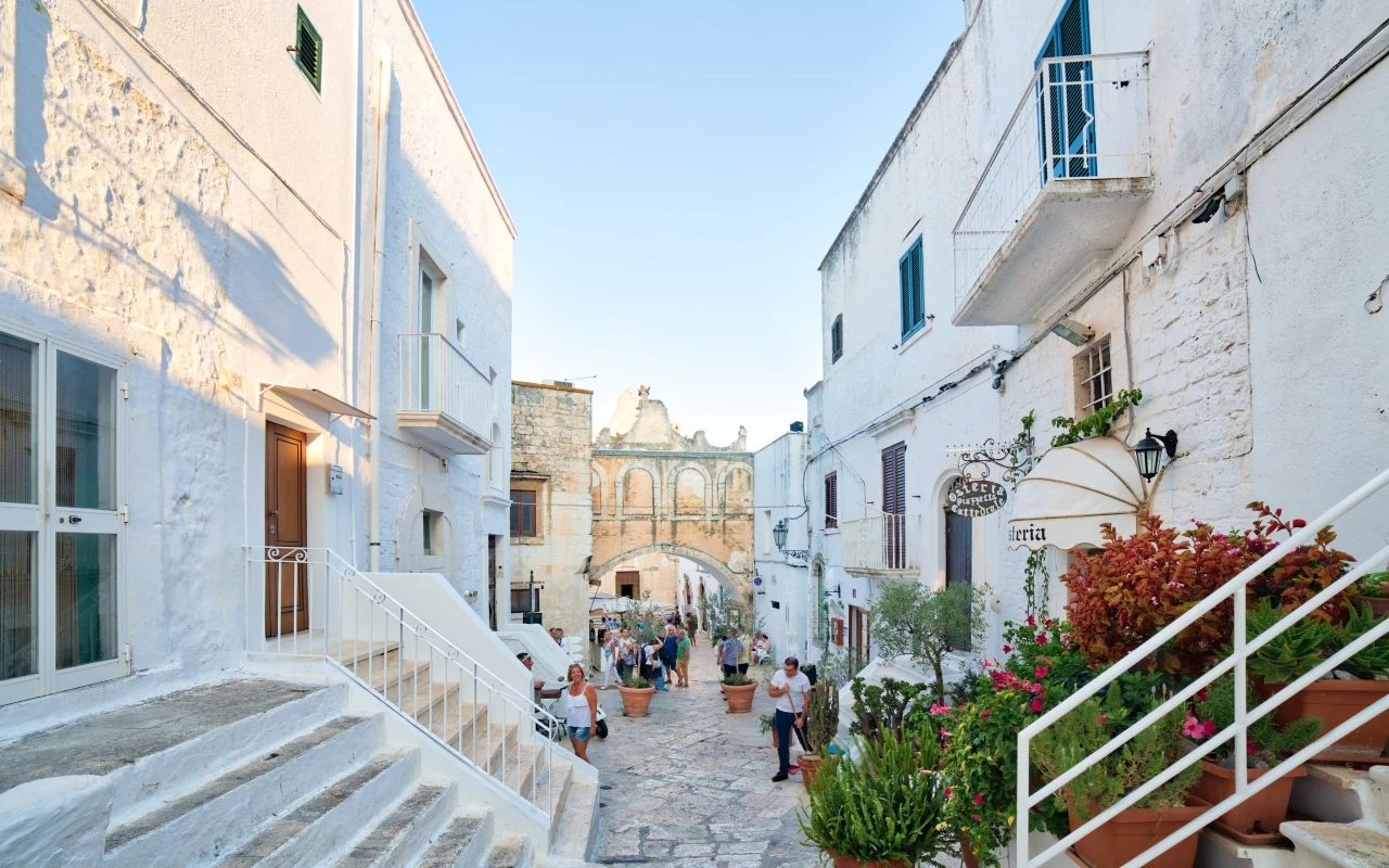 Ostuni Wąska kamienna uliczka otoczona bielonymi budynkami, liczne donice z kwiatami oraz grupy spacerujących ludzi kierujących się w stronę ozdobnej bramy na końcu ulicy w słonecznym świetle.