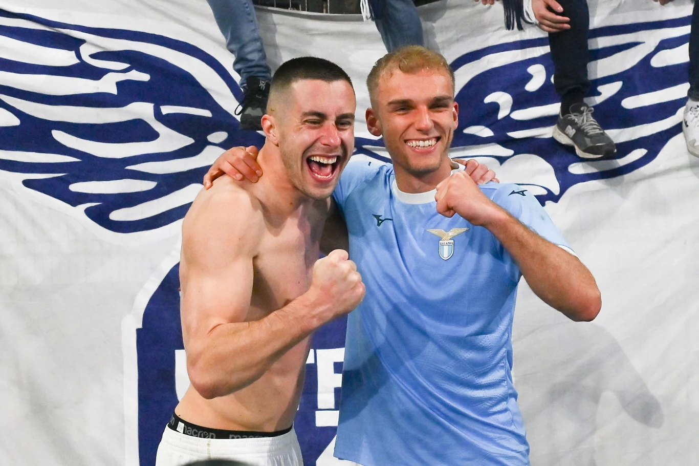 dwóch młodych mężczyzn uśmiecha się i świętuje przy wielkim kibicowskim banerze z motywem skrzydeł; jeden z nich ubrany jest w sportową koszulkę, drugi bez koszulki, obaj zaciśnięte pięści trzymają uniesione, podkreślając radość i zwycięstwo