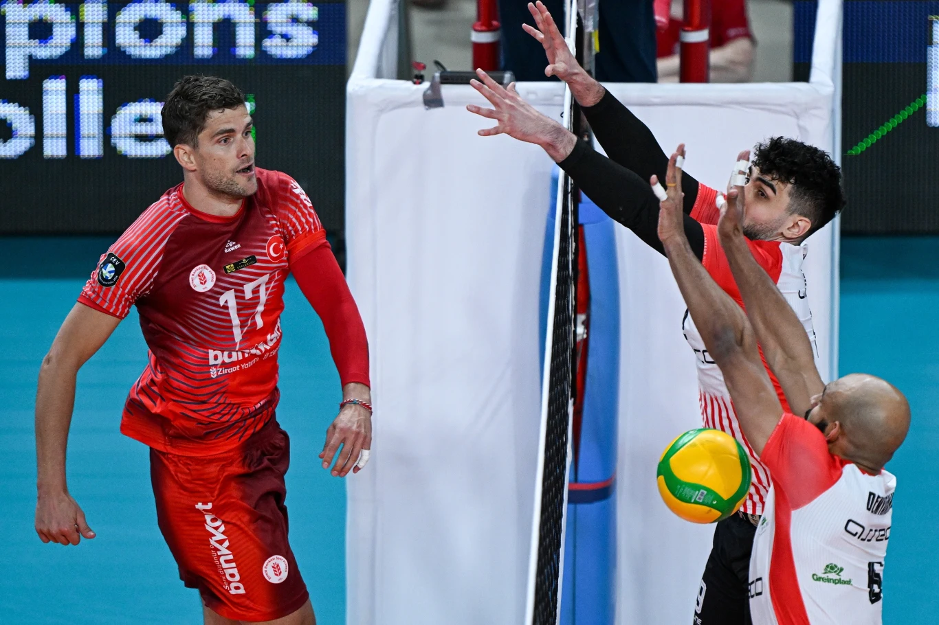 zawodnik w czerwonym stroju odbija piłkę siatkarską przy siatce, podczas gdy dwóch przeciwników w białych i czerwonych koszulkach próbuje ją zablokować