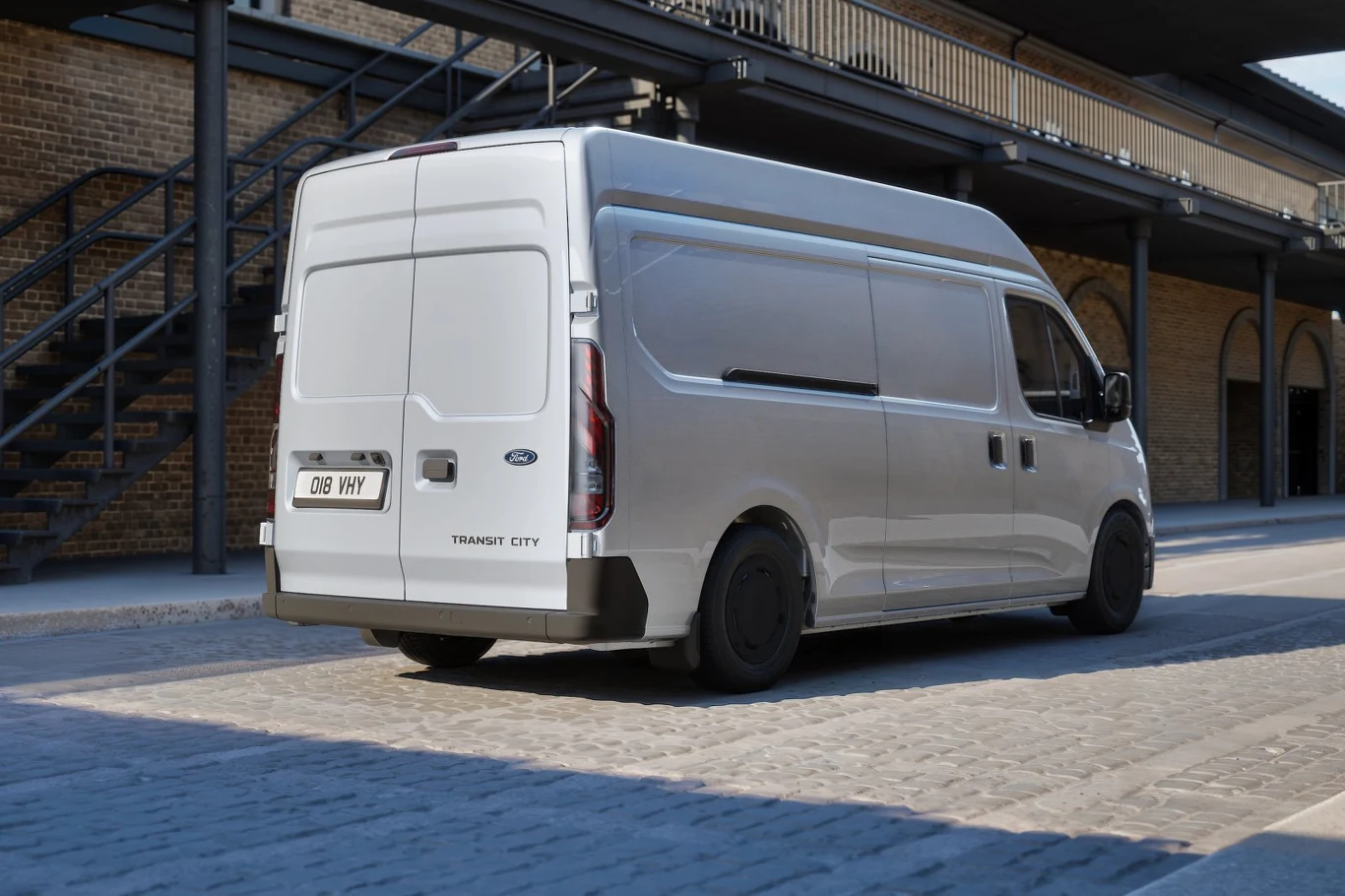 srebrny dostawczy van Ford Transit City zaparkowany na brukowanej ulicy obok budynku z metalowymi schodami i ceglaną ścianą