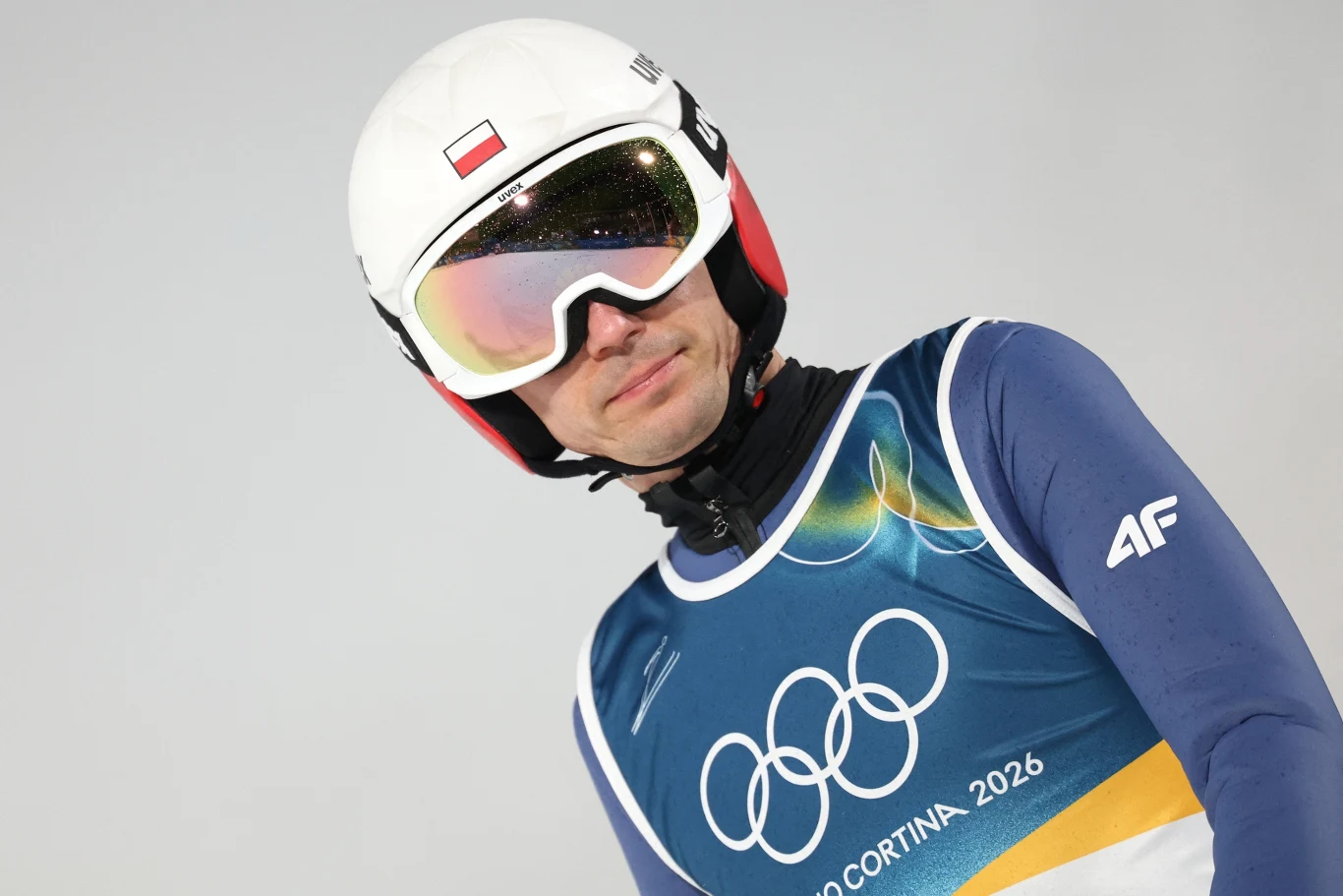 zawodnik w stroju narciarskim z kaskiem i goglami, ubrany w niebieski kombinezon z emblematem olimpijskim oraz napisem Milano Cortina 2026, na kasku widoczna flaga Polski