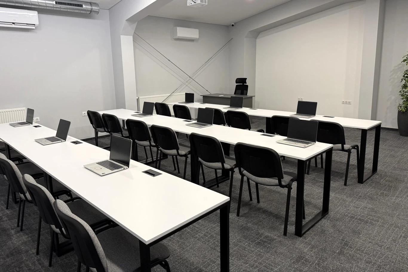 Nowoczesna sala szkoleniowa z trzema rzędami białych stołów, na których ustawione są laptopy, krzesła ustawione równo obok biurek, biurko prowadzącego na końcu sali z dużym fotelem oraz prosty, minimalistyczny wystrój z szarymi ścianami i klimatyzacją.