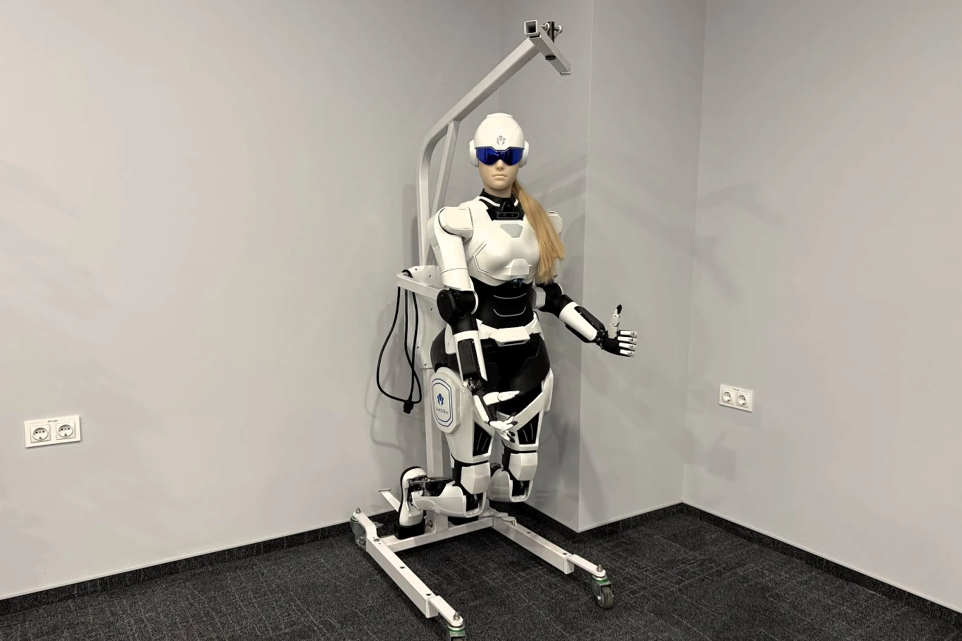 humanoidalny robot medyczny o białej i czarnej kolorystyce z blond peruką, wyposażony w okulary ochronne i zamocowany na mobilnej podstawie, ustawiony w pustym, szaro wykończonym pomieszczeniu