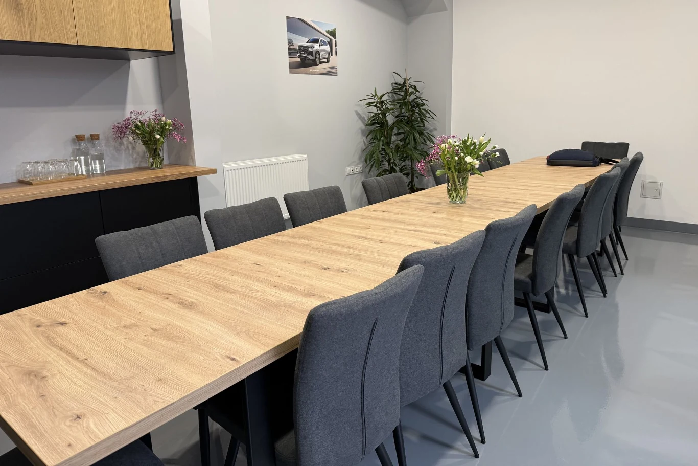 długi, drewniany stół otoczony szarymi krzesłami, na stole świeże bukiety kwiatów, w tle blat z naczyniami, roślina doniczkowa oraz obraz z motywem motoryzacyjnym na ścianie