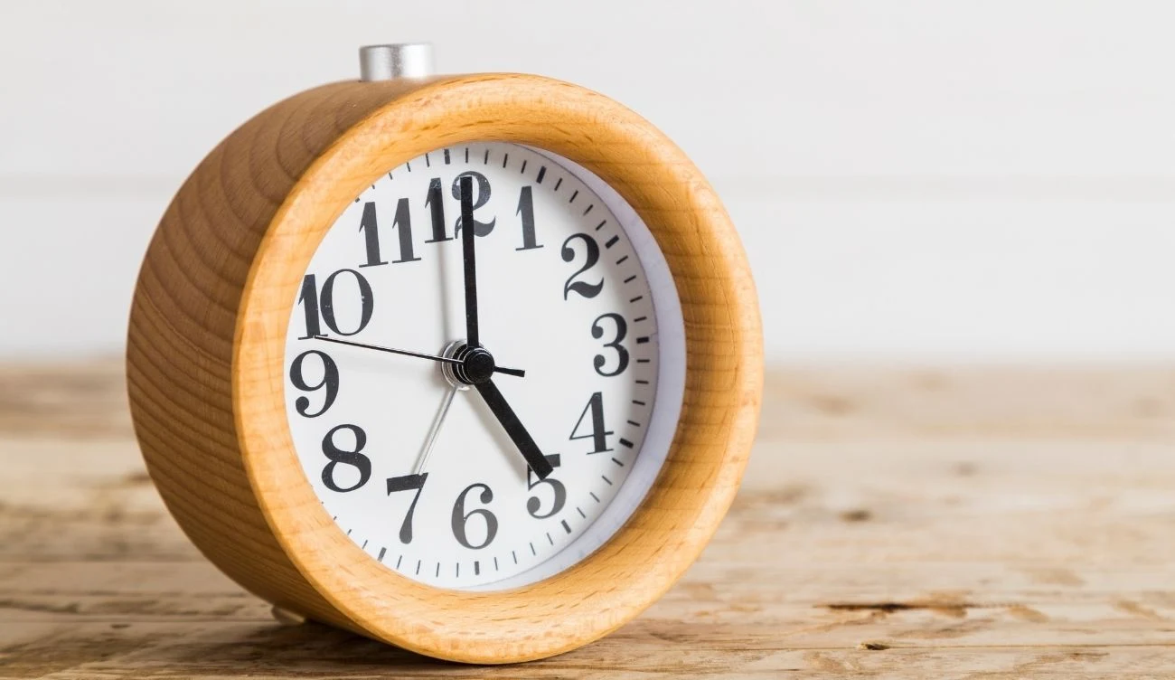 Zmiana czasu na letni ma znaczny wpływ na organizm człowieka Okrągły drewniany budzik z białą tarczą z czarnymi wskazówkami. Budzik stoi na drewnianej powierzchni, jasne tło.