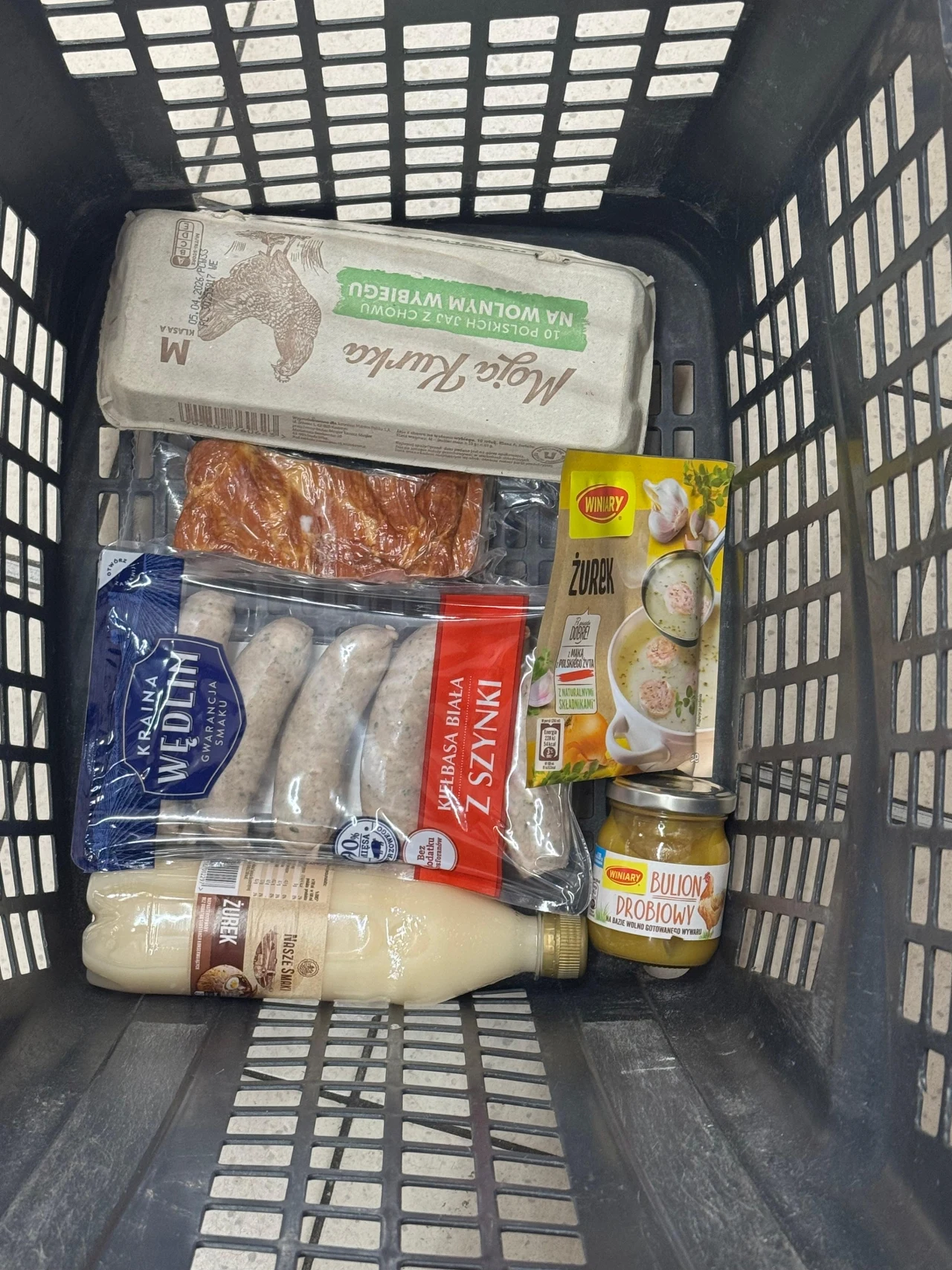 koszyk sklepowy z produktami spożywczymi: opakowanie jajek ekologicznych, paczka białej kiełbasy, opakowanie kiełbasy wędzonej, butelka białego barszczu, słoik z białą kiełbasą drobiową, opakowanie żurku w proszku oraz paczka kabanosów