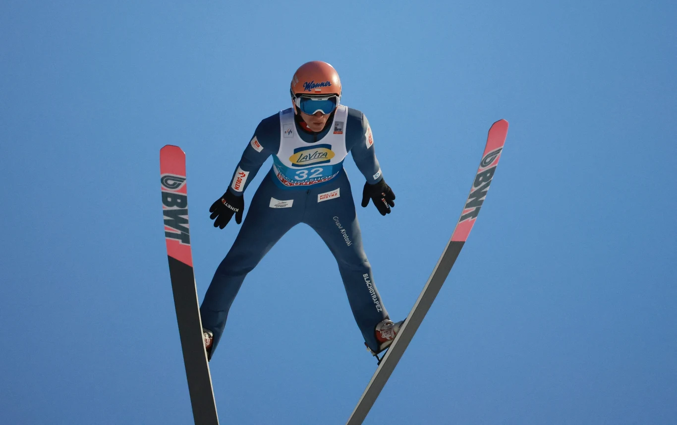 skoczek narciarski w locie z szeroko rozstawionymi nartami na tle czystego, niebieskiego nieba, ubrany w niebieski kombinezon i kask, z widocznym numerem startowym oraz oznaczeniami sponsorskimi na nartach