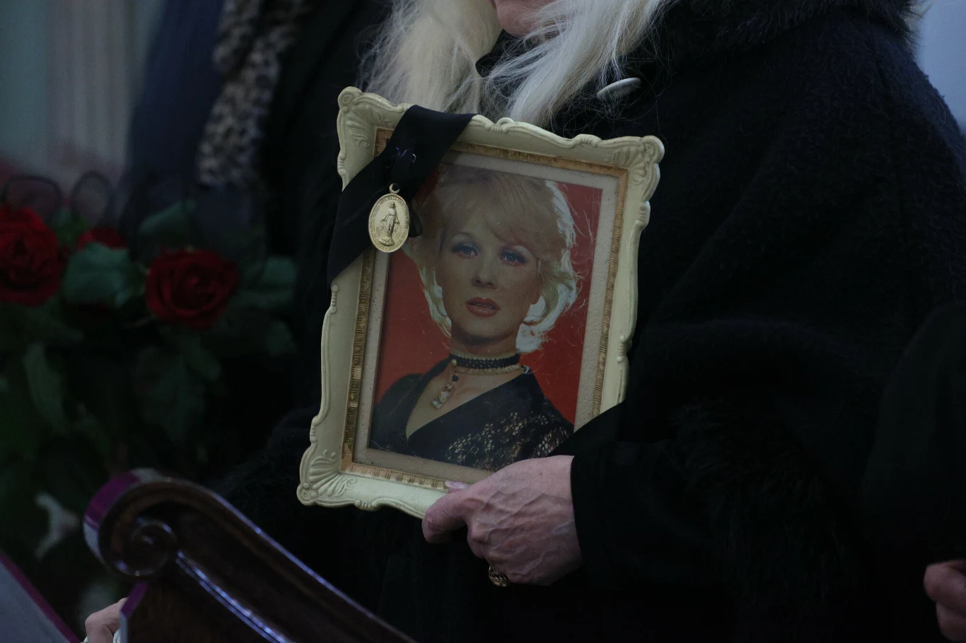 osoba ubrana na czarno trzyma w rękach portret kobiety w ozdobnej ramce, do której przypięta jest czarna wstążka z medalionem; w tle widoczne czerwone róże