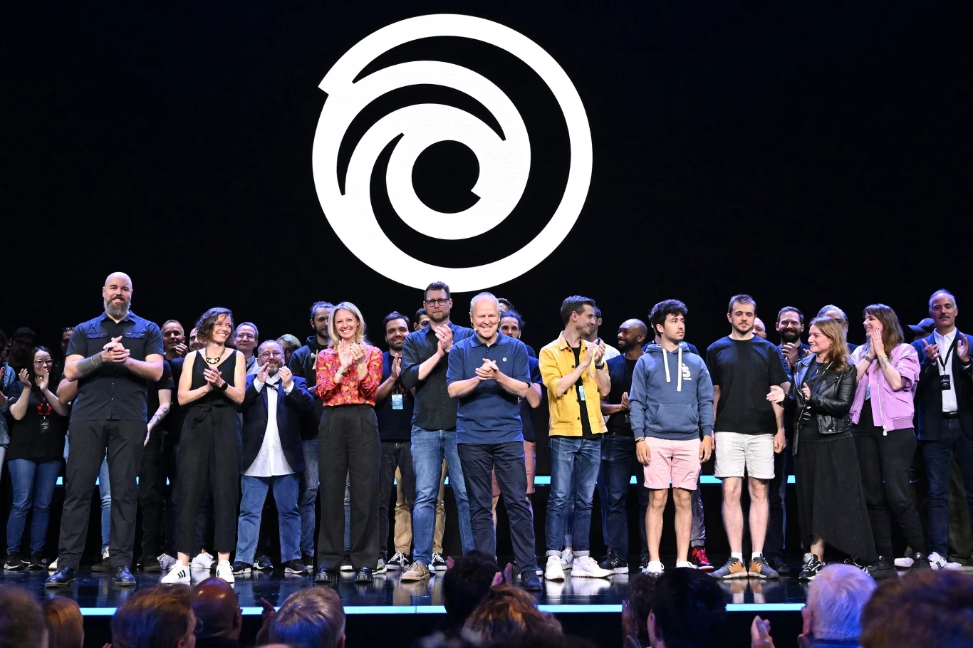 Grupa ludzi stojąca na scenie, wielu z nich bije brawo, za nimi duże logo Ubisoft, scena oświetlona, tłum widzów w części widoczny na pierwszym planie.