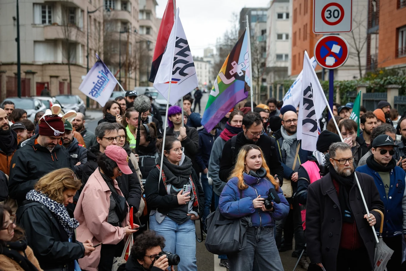 grupa ludzi zgromadzona na ulicy miasta, wiele osób trzyma flagi i transparenty, na pierwszym planie widoczne są osoby z aparatami fotograficznymi oraz w zimowych ubraniach, w tle znajdują się wielopiętrowe budynki mieszkalne oraz znaki drogowe