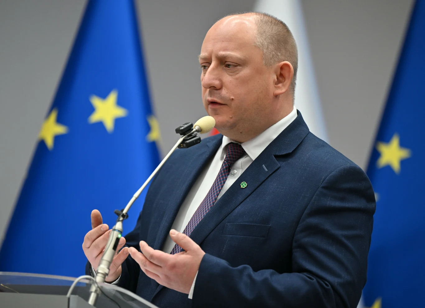 mężczyzna w garniturze mówi do mikrofonu, gestykuluje dłońmi, w tle flagi Unii Europejskiej i Polski
