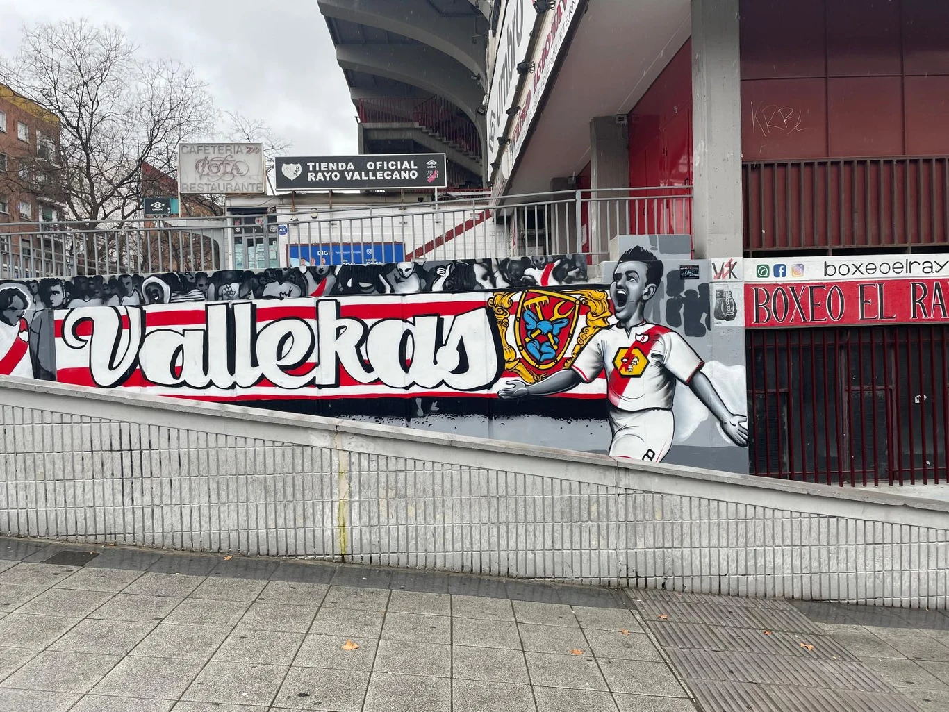 Kolorowy mural z napisem 'Vallekas', herbem klubu Rayo Vallecano oraz wizerunkiem piłkarza w biało-czerwonym stroju z uniesionymi rękami w geście radości; w tle widoczne sylwetki kibiców oraz fragment stadionu i sklepu klubowego.