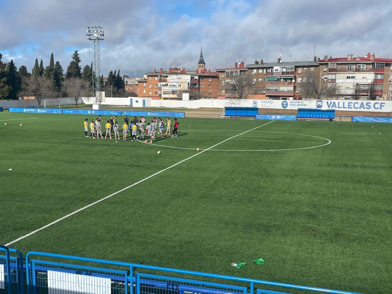 grupa piłkarzy w treningowych strojach zgromadzona w kręgu na środku boiska piłkarskiego, otoczonego niebieskimi barierkami i zabudowaniami w tle, z wyraźnym napisem 'Vallecas CF' na bandzie