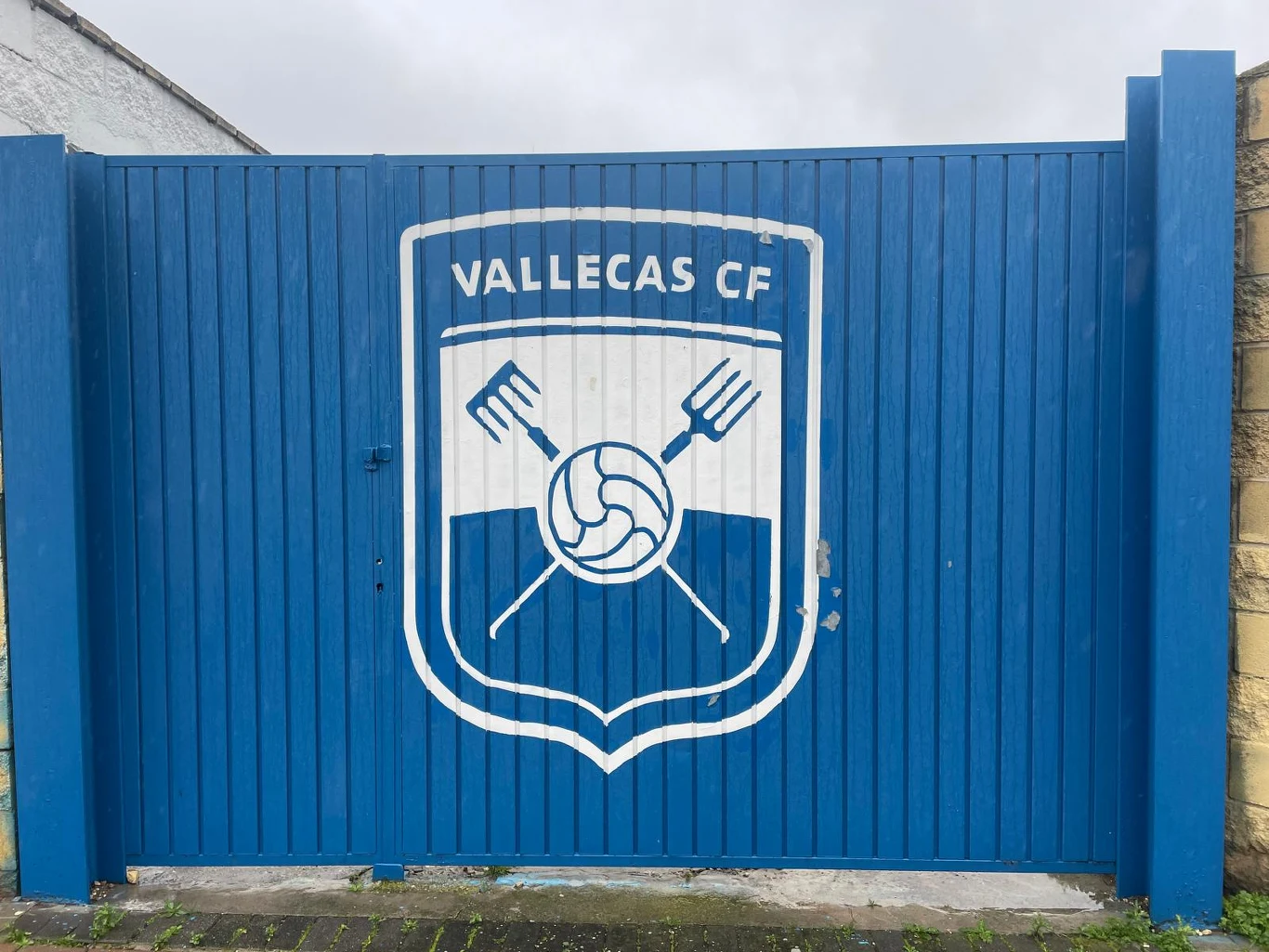 niebieska brama z dużym herbem klubu Vallecas CF, na którym umieszczono piłkę oraz dwa skrzyżowane widły po bokach