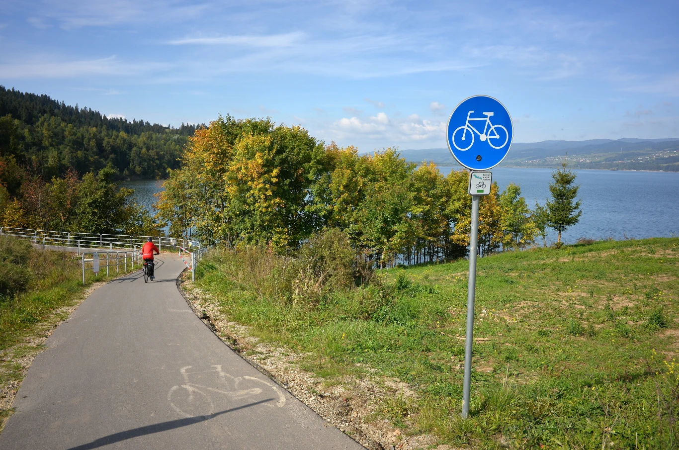 asfaltowa ścieżka rowerowa prowadząca w kierunku jeziora, po lewej stronie rowerzysta w czerwonej kurtce, obok niebieski znak szlaku rowerowego, w tle drzewa oraz widoczna tafla wody