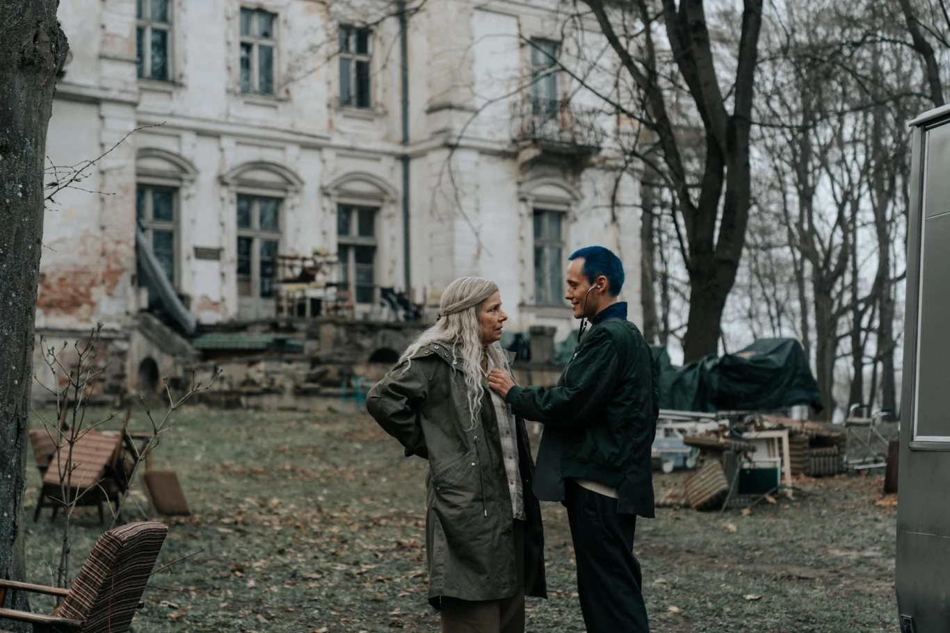 Dwie osoby stoją naprzeciw siebie na podwórzu opuszczonego, zniszczonego budynku, otoczonego przez gołe drzewa i stare, rozrzucone meble ogrodowe; atmosfera sceny jest poważna i nieco ponura.