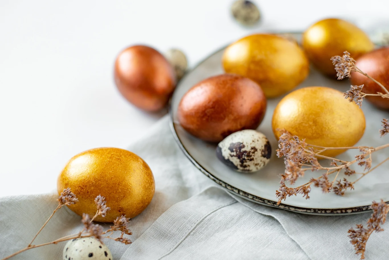 Ta przyprawa zabarwi pisanki na piękny, złocisty kolor Pisanki barwione kurkumą. Jajka w odcieniach złota i miedzi na szarej serwetce i talerzu, ozdobione suchymi gałązkami roślin