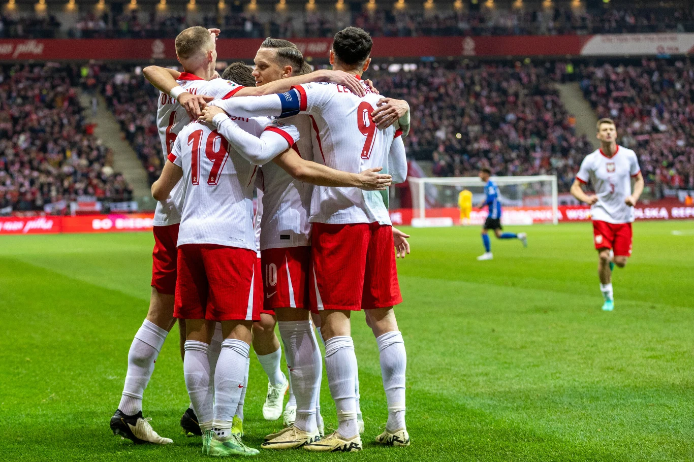 piłkarze polskiej reprezentacji w biało-czerwonych strojach świętują strzelenie gola, otoczeni przez kibiców na trybunach, jeden zawodnik z niebieskiej drużyny widoczny w tle