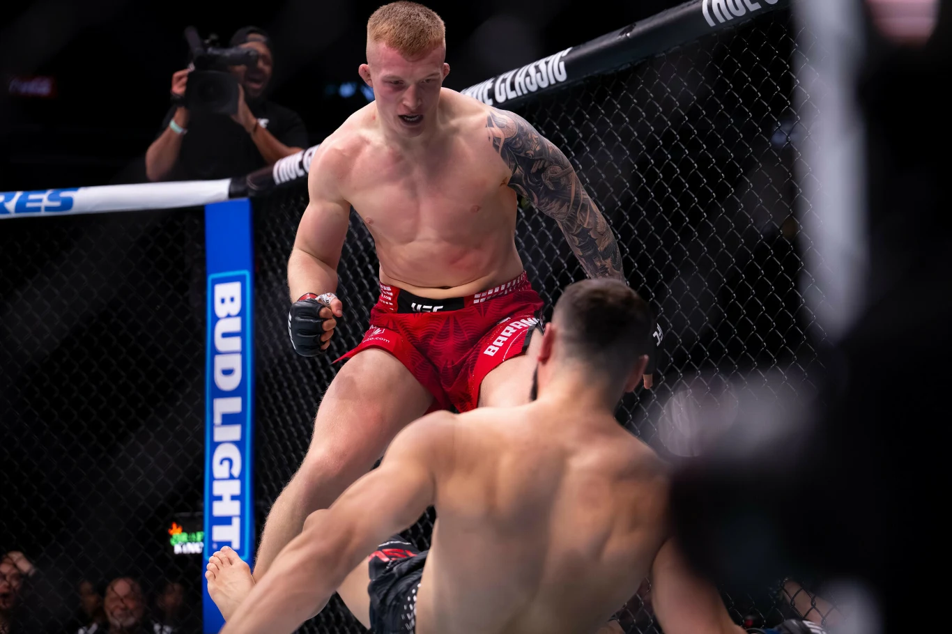 zawodnik MMA w czerwonych spodenkach atakuje przeciwnika, który upada na matę klatki, obok znajdują się siatka oktagonu oraz wyraźne logo sponsora