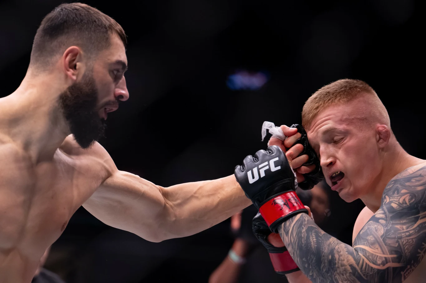 dwóch zawodników MMA w trakcie walki, jeden trafia przeciwnika pięścią w twarz, a drugi z zamkniętymi oczami przyjmuje cios, obaj ubrani w rękawice i sportowe spodenki, w tle zaciemniona arena