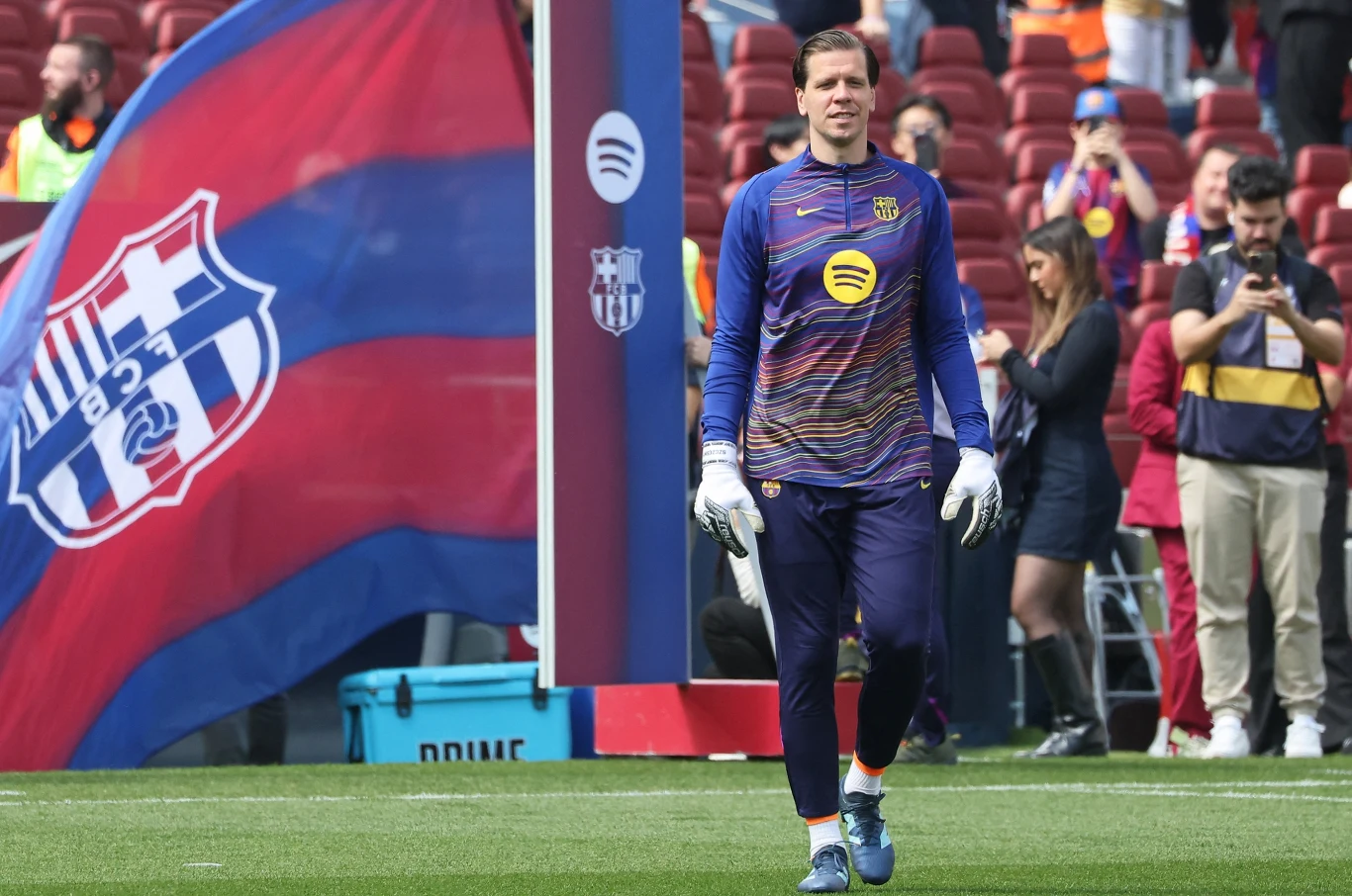 bramkarz w stroju treningowym z logotypem sponsora, stojący na murawie stadionu piłkarskiego, w tle duża flaga FC Barcelona oraz trybuny z kibicami