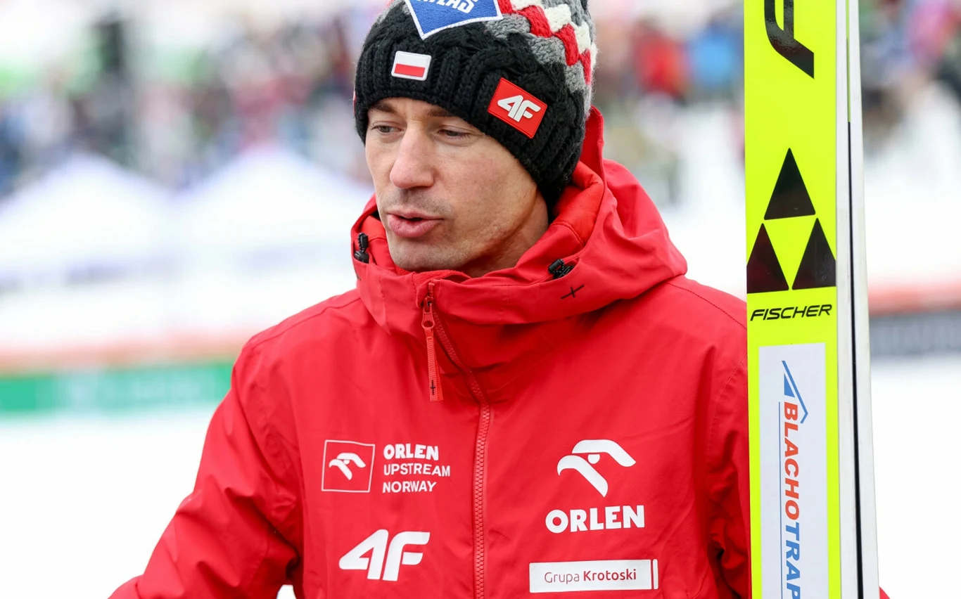 mężczyzna w czerwonej kurtce sportowej z logo Orlen i czapce z polską flagą trzyma narty marki Fischer, w tle rozmazany biały krajobraz zimowy