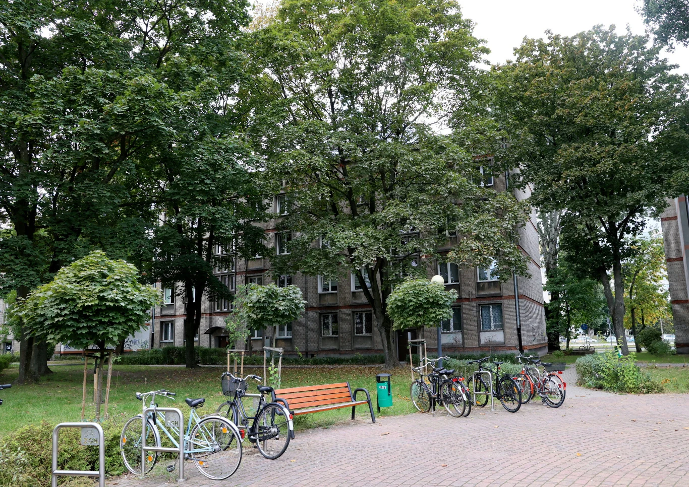 ławkę ustawioną wśród zielonych drzew i kilku stojaków rowerowych z przypiętymi rowerami, w tle widać szare budynki mieszkalne z licznymi oknami