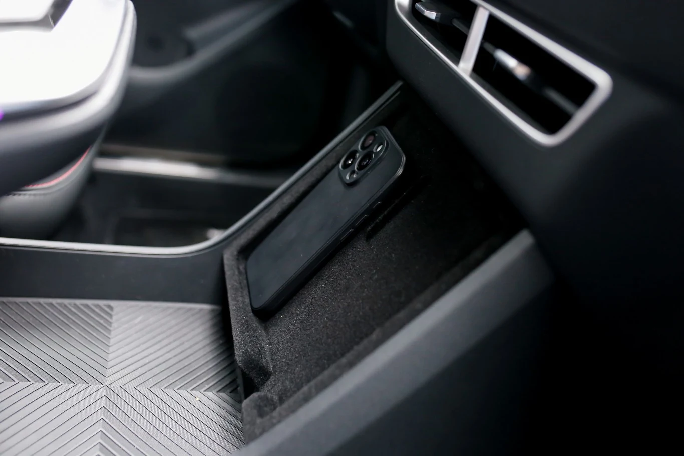 smartfon w czarnym etui umieszczony w przeznaczonym na to schowku w konsoli środkowej samochodu, obok widoczny fragment nowoczesnego wnętrza i kratki wentylacyjnej
