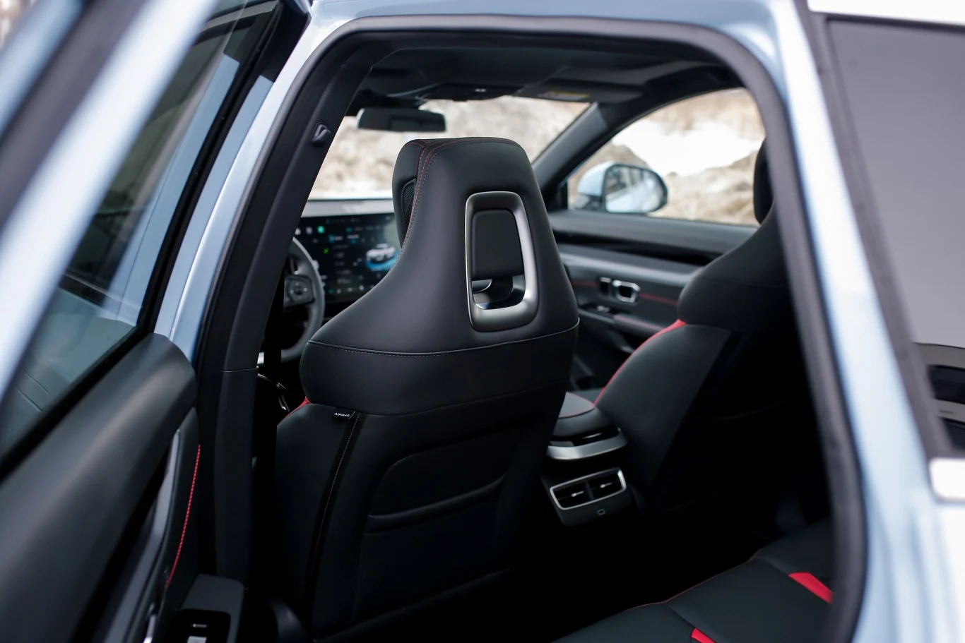 widok wnętrza samochodu z otwartymi tylnymi drzwiami, nowoczesny fotel kierowcy z metalicznym otworem zagłówka, nowoczesna deska rozdzielcza z dużym ekranem dotykowym i kontrastującymi czerwonymi przeszyciami na boczkach foteli