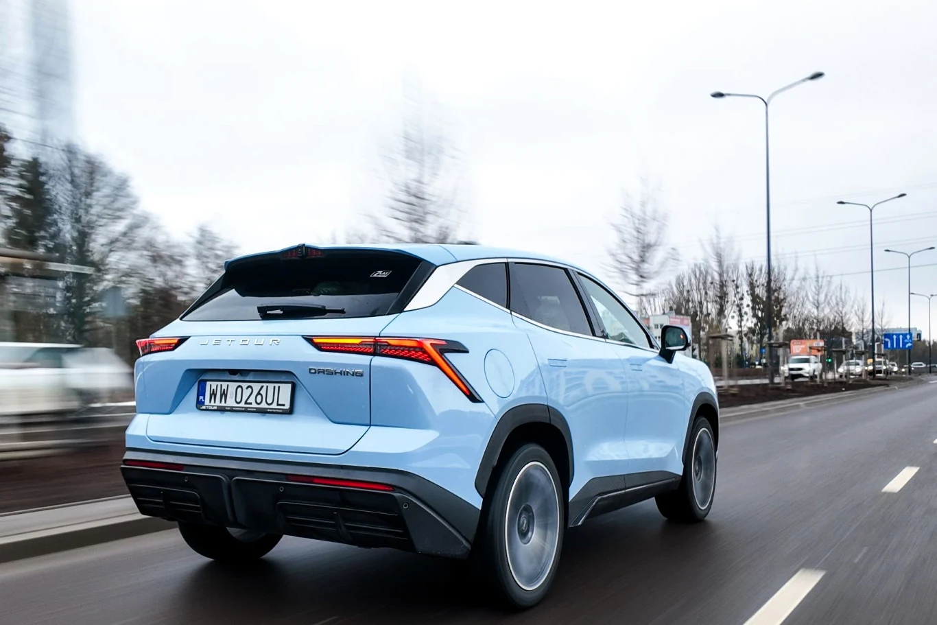 jasnoniebieski SUV z nowoczesnym designem i ostrą linią tylnych świateł porusza się po miejskiej ulicy, otoczony nagimi drzewami i ulicznymi latarniami