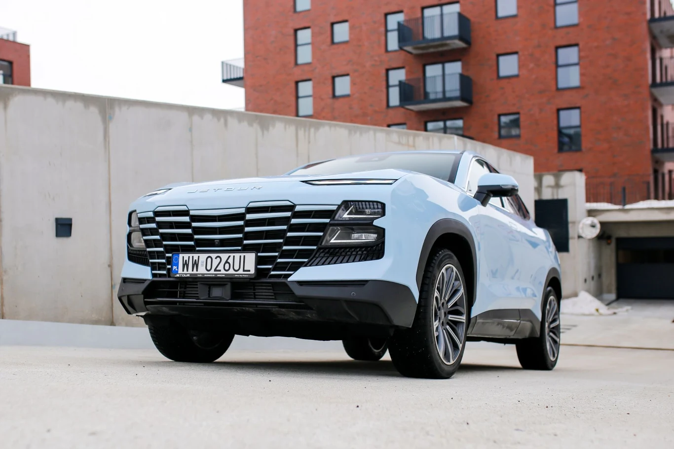 jasnoniebieski samochód SUV o nowoczesnym kształcie, z charakterystycznym czarnym grillem i reflektorami LED, zaparkowany na tle nowoczesnych budynków mieszkalnych z czerwonej cegły