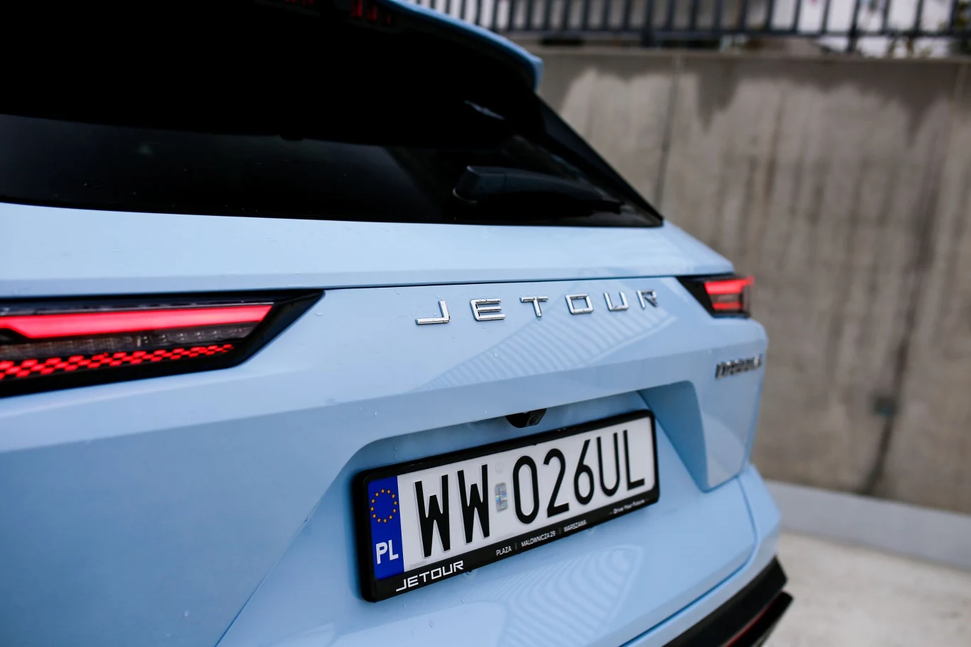 tylny fragment jasnoniebieskiego samochodu marki Jetour z wyraźnie widocznymi czerwonymi lampami LED i polską tablicą rejestracyjną WW 026UL; w tle betonowy mur