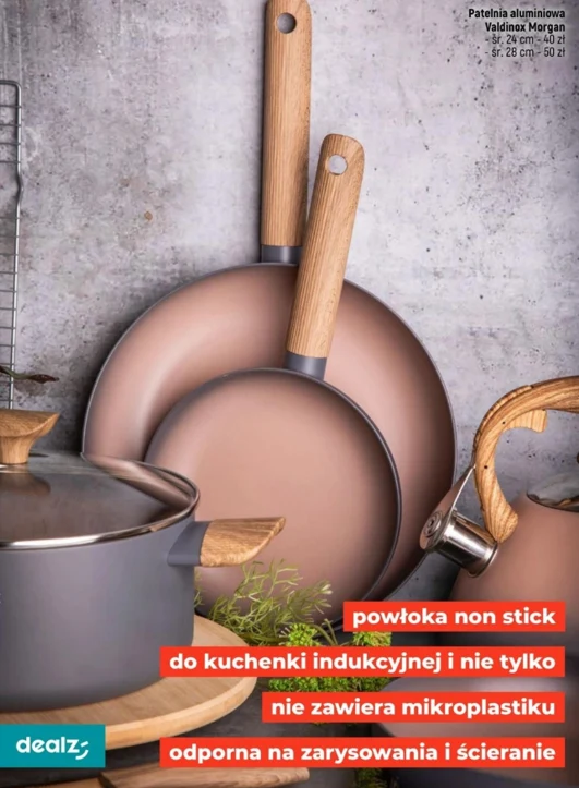 zestaw aluminiowych patelni i garnków z drewnianymi rączkami oraz powłoką non stick, ustawionych na tle betonowej ściany, z widocznymi etykietami informującymi o ich odporności na zarysowania, uniwersalności oraz braku mikroplastiku