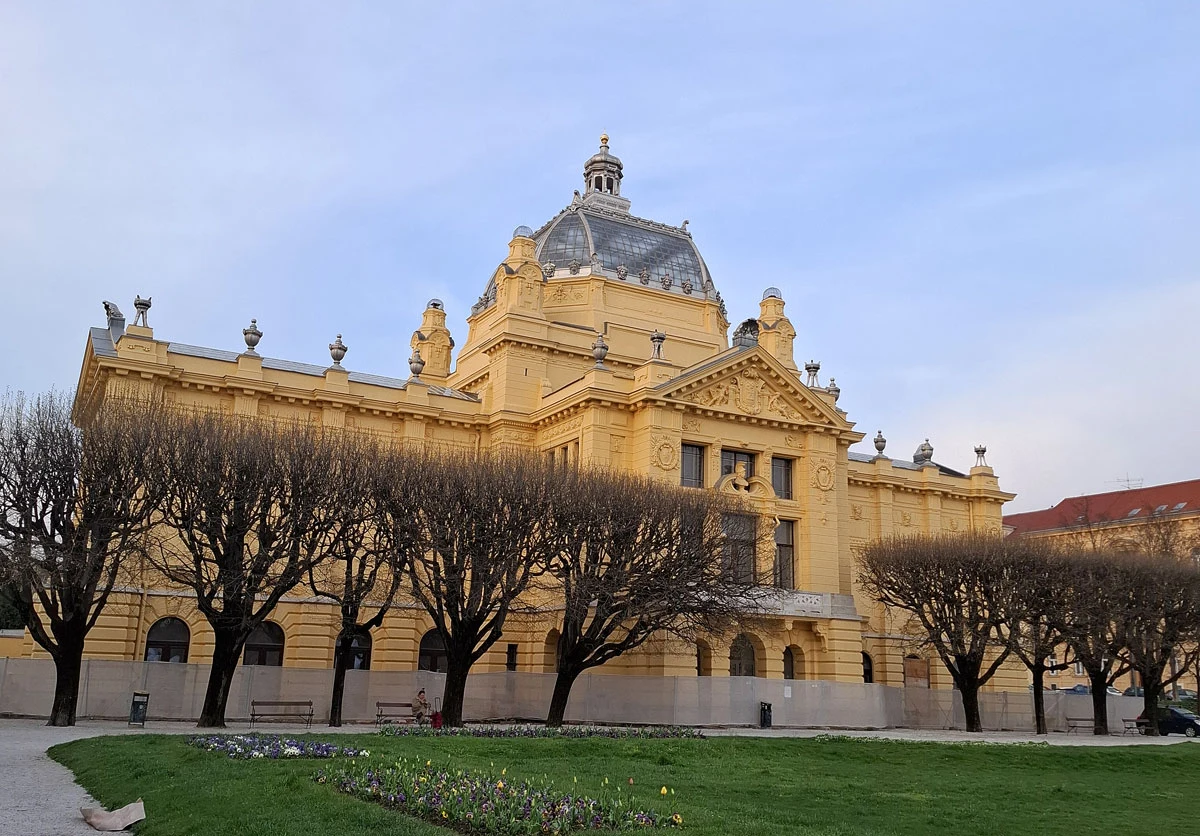 Żółta, zabytkowa hala wystawiennicza z dużą kopułą, ozdobnymi rzeźbami i kolumnami, otoczona przyciętymi, bezlistnymi drzewami oraz trawnikiem z kwiatową rabatą na pierwszym planie.