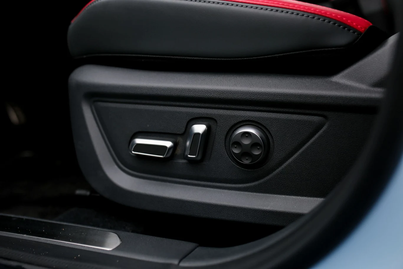 panel sterowania elektrycznym fotelem samochodowym z widocznymi przyciskami regulacji pozycji oraz okrągłym pokrętłem, obramowany czarną skórą z czerwonym wykończeniem