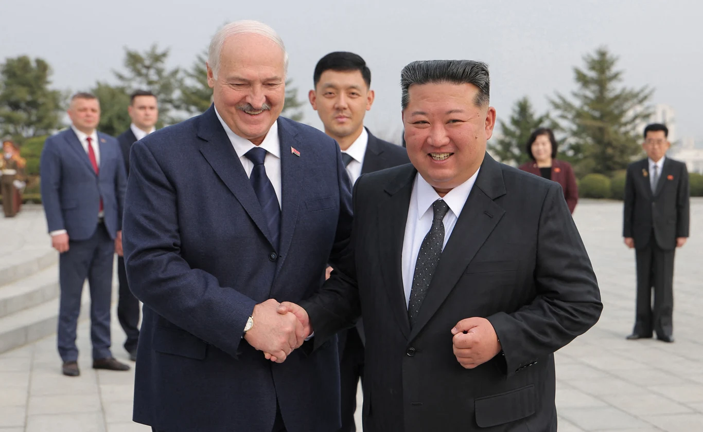 Dwóch mężczyzn w ciemnych garniturach uśmiecha się i ściska dłonie, w tle kilka osób formalnie ubranych stoi na brukowanym placu z drzewami ozdobnymi.