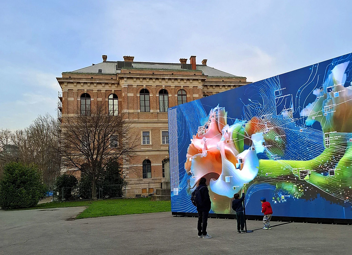 trzy osoby stoją przed dużym, kolorowym muralem z abstrakcyjną grafiką cyfrową, a w tle widać zabytkowy budynek z cegły