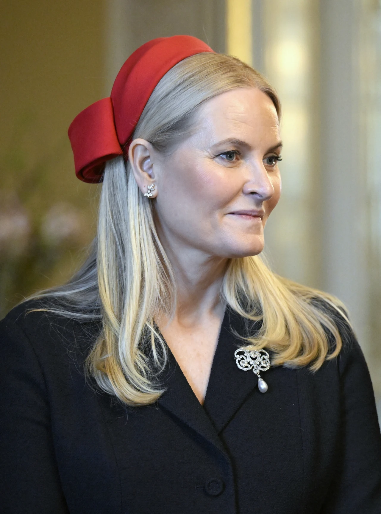 Kobieta o jasnych, prostych włosach, ubrana w elegancki czarny żakiet z ozdobną broszką oraz perłowymi kolczykami, na głowie ma efektowną, dużą czerwoną kokardę jako opaskę.