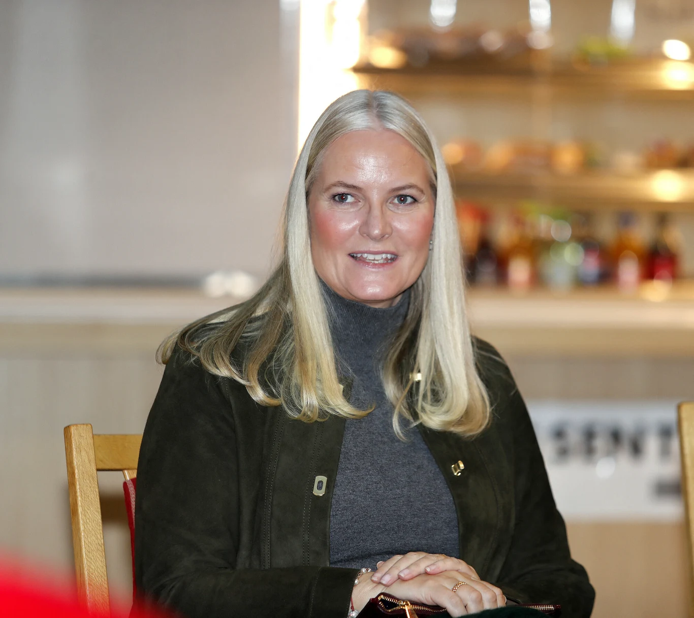 kobieta o jasnych, prostych włosach siedzi na krześle, ubrana w ciemną marynarkę i szary golf, w tle rozmyta półka z napojami oraz jedzeniem