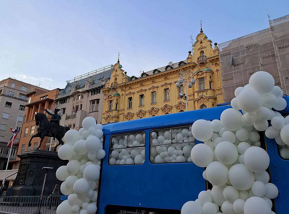 niebieski autobus wypełniony białymi balonami otoczony kolejnymi balonami, w tle historyczna żółta kamienica i pomnik konny na placu miejskim